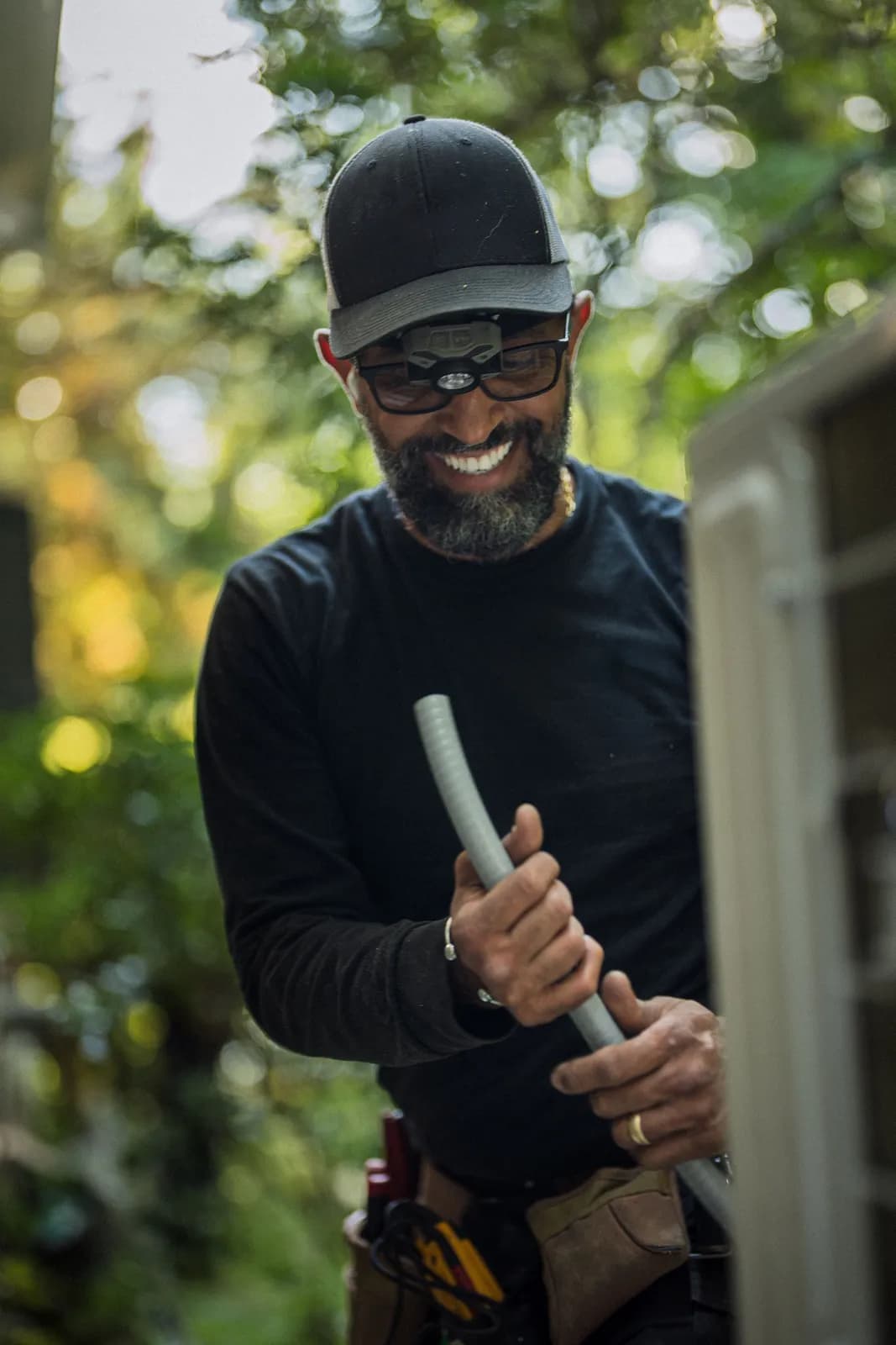 Smiling electrician in a work cap and tool belt handling flexible conduit outside a home
