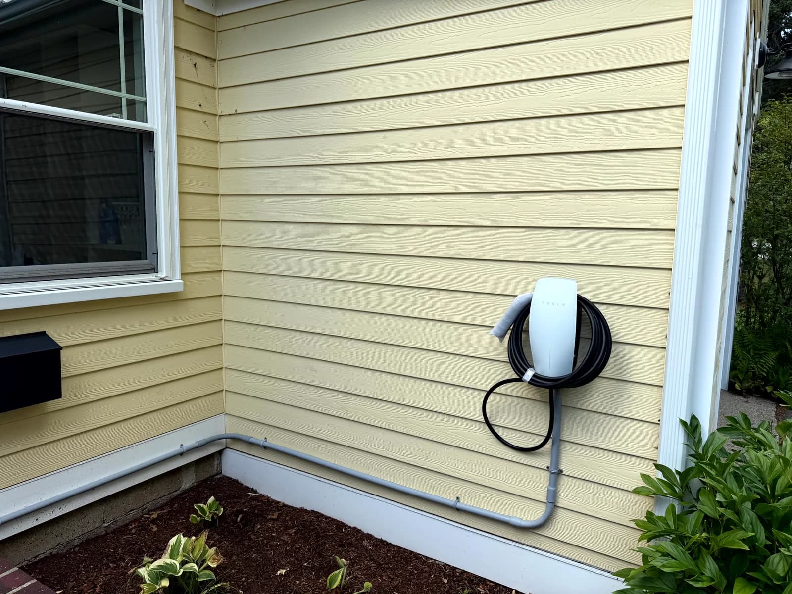 Tesla Wall Connector mounted on the exterior siding of a home with conduit run along the foundation